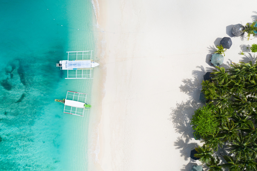 Aerial view of two traditional outrigger boats moored in shallow turquoise water off a white sand beach with mangrove vegetation and palm trees along the shoreline.