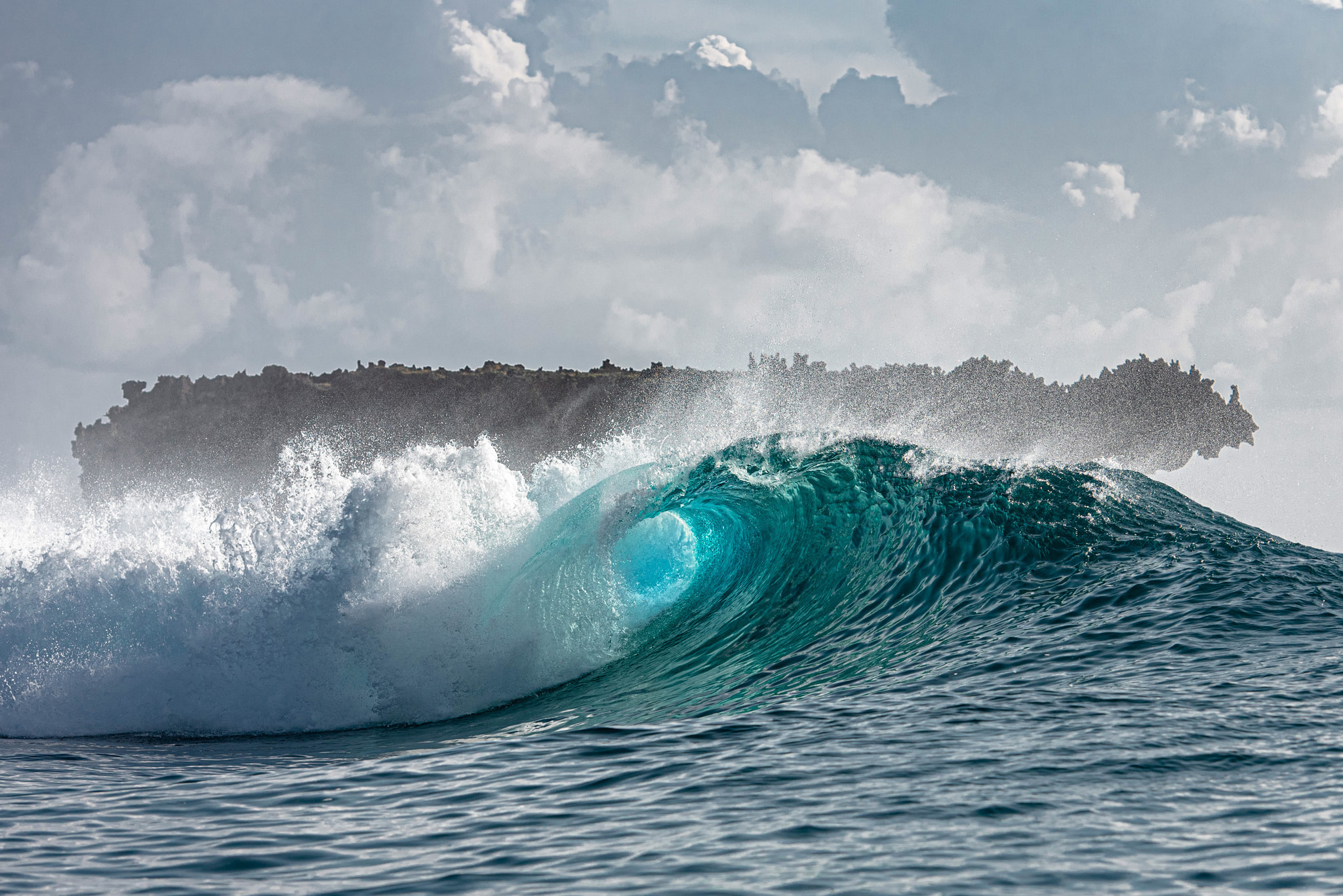 A powerful wave with a turquoise barrel and white crest breaks offshore under a partly cloudy sky.