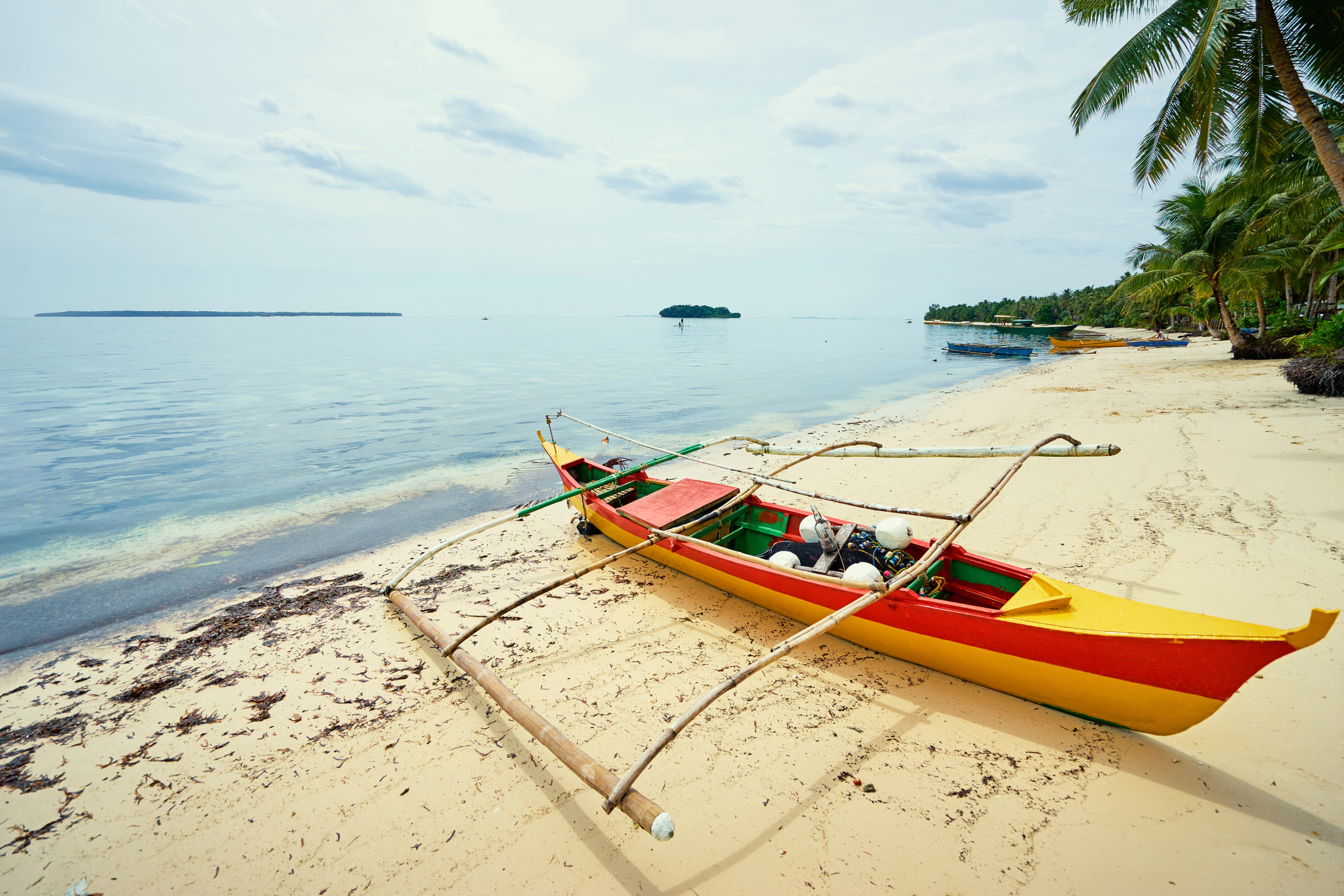 A traditional red and yellow outrigger boat rests on the sand of a pristine tropical beach, with calm turquoise water and palm-fringed shoreline beyond.
