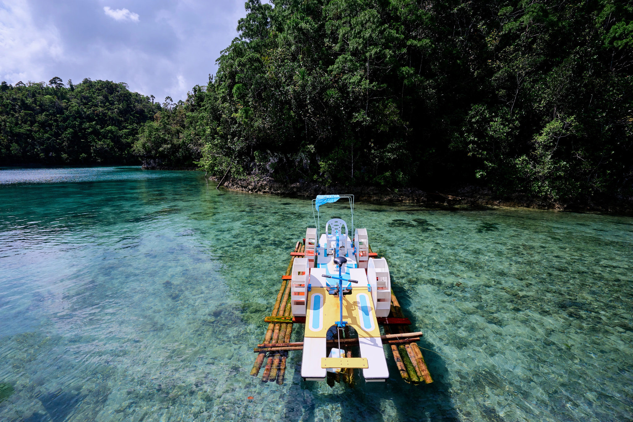 A traditional wooden outrigger boat moored in crystal-clear turquoise lagoon water with dense mangrove and tropical forest lining the shore.
